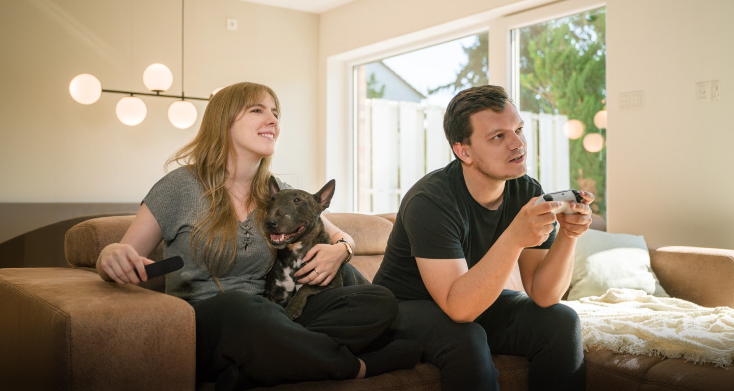 Un couple sur un canapé, la femme avec une télécommande et un chien, l'homme jouant à un jeu vidéo.