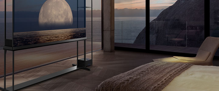  LG SIGNATURE OLED T displays a moon depicted above a shore on the transparent screen. The LG SIGNATURE OLED T sits in the middle of a room with a cliff side view that looks out to the sea. On the right is a bed and a lounge chair. 