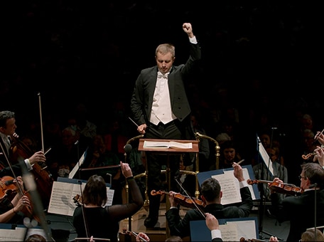 A conductor directs an orchestra. Overlayed text: 'The Musical Offering to the Whole World. #music #partnership'