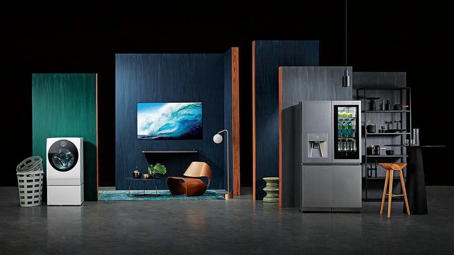 starting from the left, lg signature washing machine, oled tv w, refigerator is displayed with some green and blue wall in some place like a dark studio