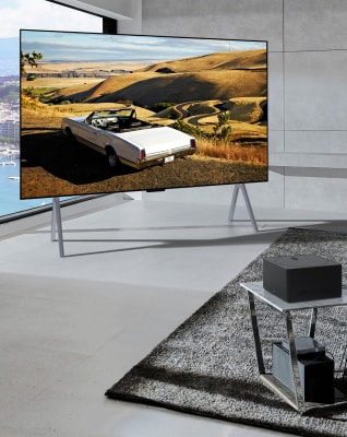 In front of a window with a view of the sea, there is a 97-inch OLED M TV placed, and in front of that, there is a table with a zero connect box on top.