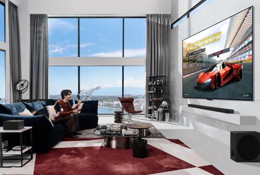 In a stylish living room overlooking the sea, a man is enjoying a car racing game on OLED M TV with zero connect box.