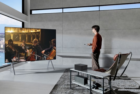 On the table, there is a zero connect box placed, below which sits a speaker, next to which a man stands, enjoying an orchestra performance on OLED M TV.