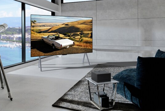 In front of a window with a view of the sea, there is a 97-inch OLED M TV placed, and in front of that, there is a table with a zero connect box on top.