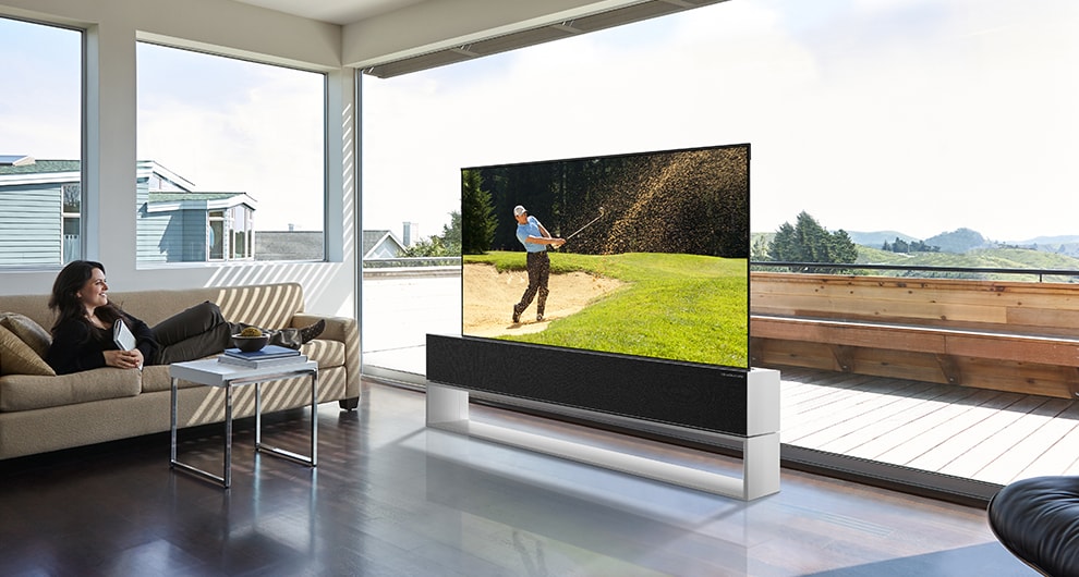 Eine Frau, die auf einem Sofa sitzt, sieht sich in ihrem geräumigen Wohnzimmer ein Golfturnier auf ihrem rollbaren OLED-Fernseher LG SIGNATURE an.