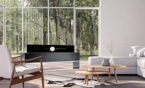 Ein Mann lehnt sich gegen ein großes Fenster, um die Nachtansicht der Stadt zu sehen, und neben ihm steht der LG SIGNATURE Rollale OLED-Fernseher in Linienansicht.