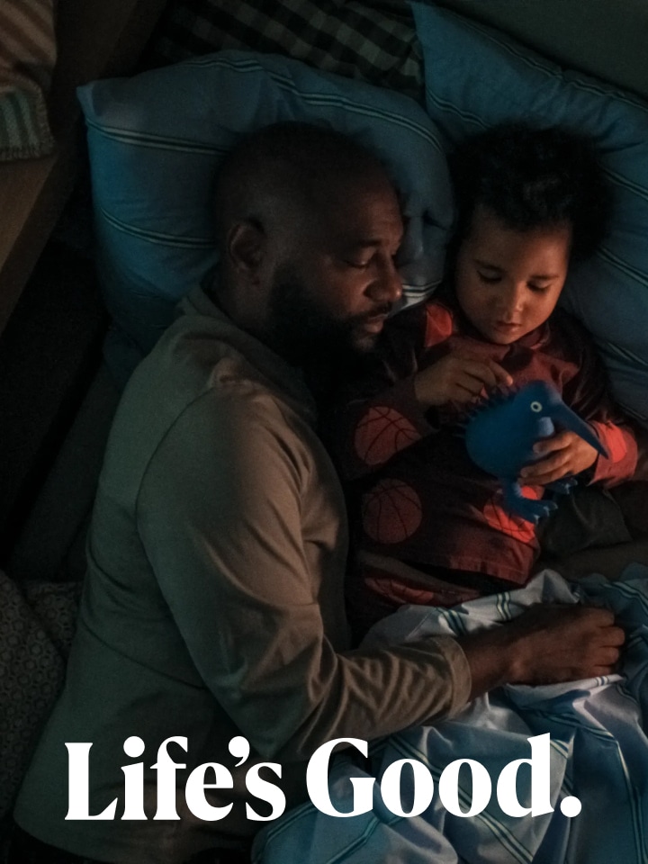 A child playing with a blue toy on a bed in their father's arms, with the small text "Life's Good" below.