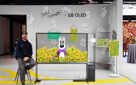 Artist Steven Harrington is sitting on a stool beside a transparent LG OLED TV that displays his signature artwork in a themed exhibition space.