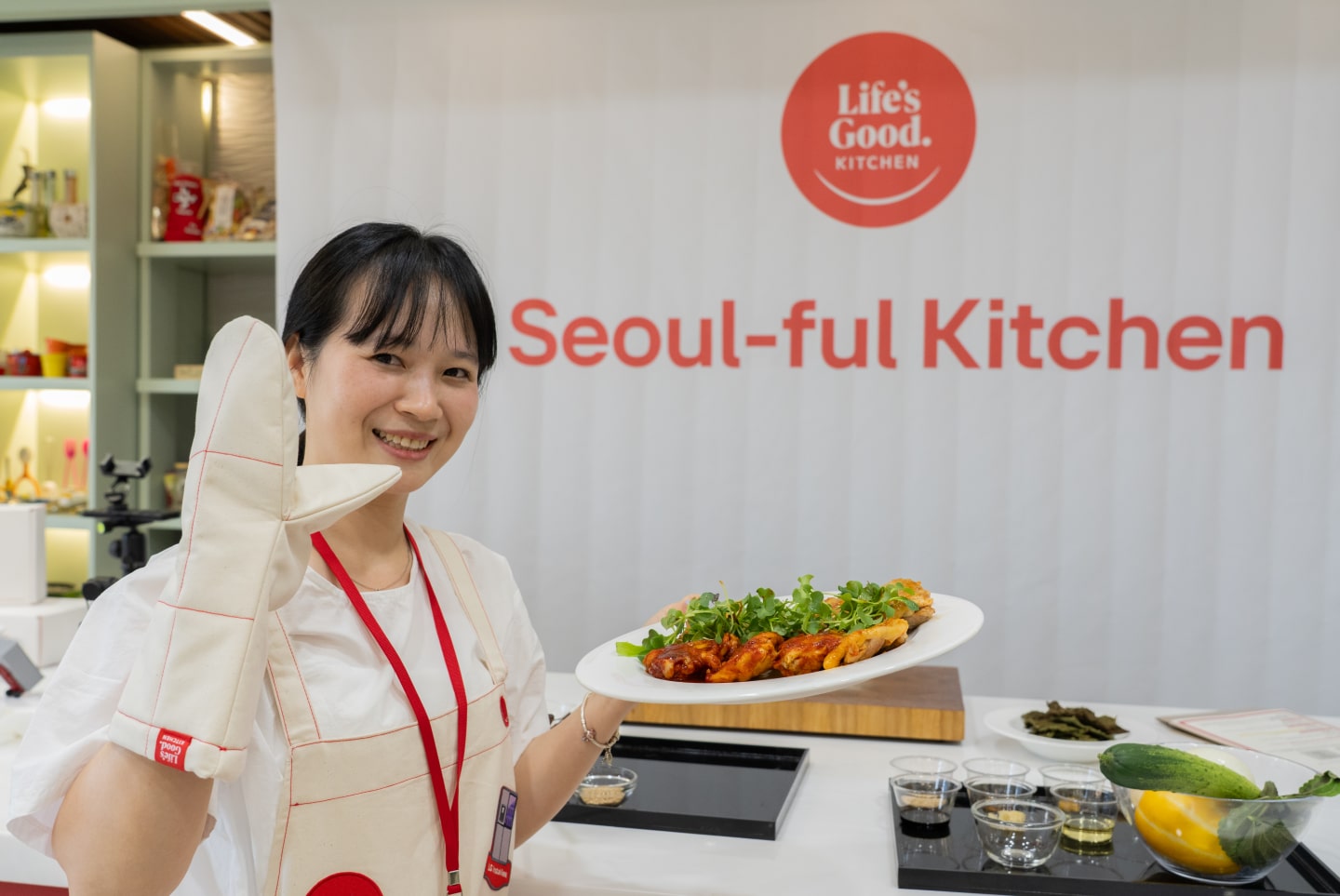A woman wearing an oven mitt waves and holds a platter of food in front of a “Seoul-ful Kitchen” backdrop at LG’s Seoul-ful Kitchen Challenge experience space.