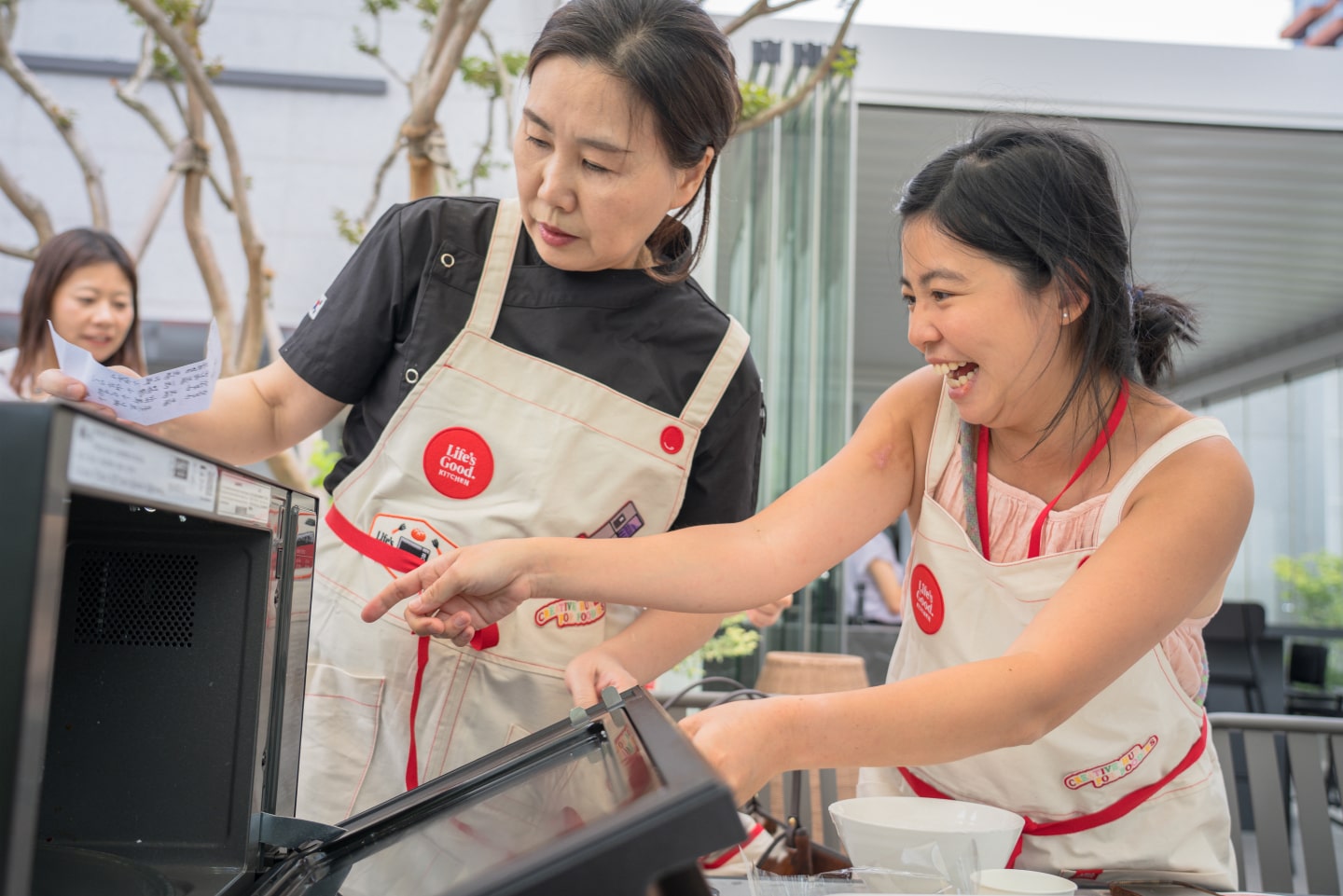 Two women wearing aprons interact with an LG oven, smiling and pointing at the appliance in an outdoor cooking area.