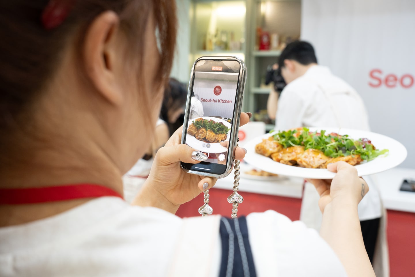 A person takes a photo of a plated dish with a smartphone, with the Seoul-ful Kitchen Challenge experience space visible in the background.