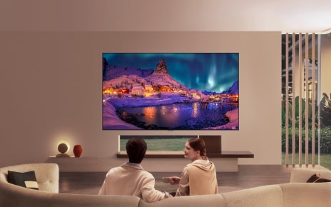 LG OLED evo is flush-fit wall-mounted in a modern living room, displaying a vivid aurora over a snowy coastal village while a man and woman watch from the sofa.