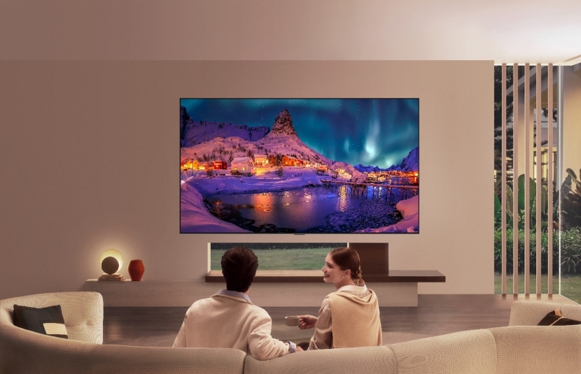 LG OLED evo is flush-fit wall-mounted in a modern living room, displaying a vivid aurora over a snowy coastal village while a man and woman watch from the sofa.