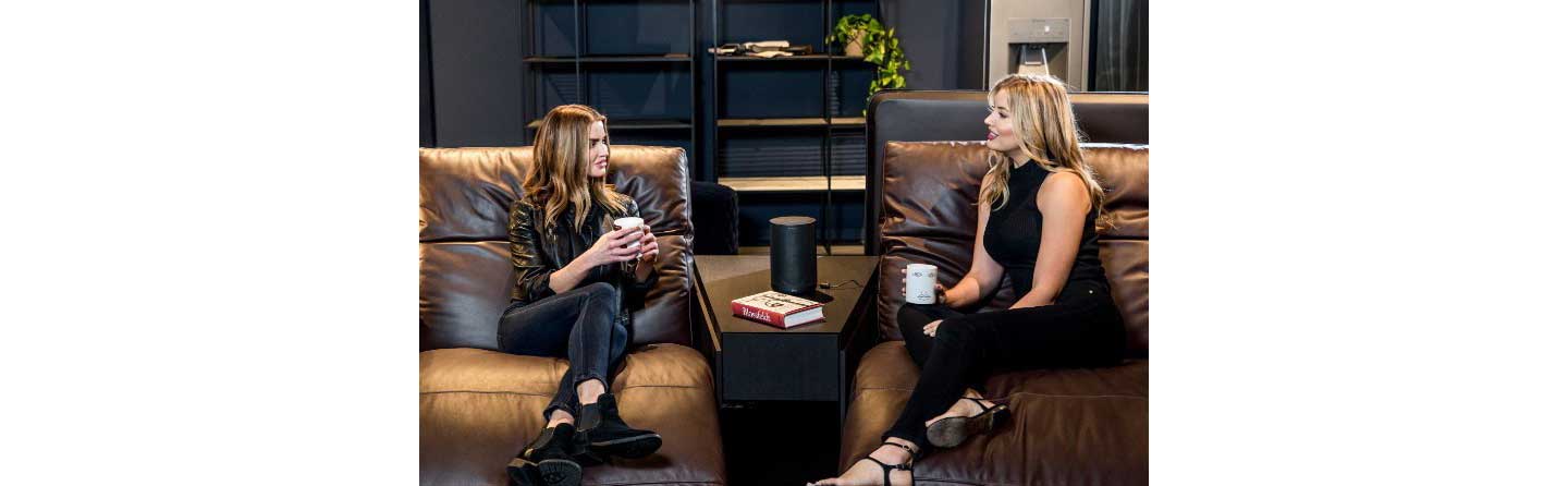 Two women sit on Natuzzi’s Colosseo sofa, talking to each other with LG’s AI smart speaker placed between them.