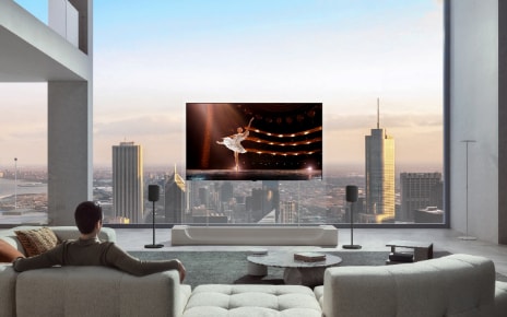 A man is sitting on a sofa in his city apartment, watching a ballerina performance on an LG OLED evo AI W6 Wallpaper TV mounted on a transparent stand with two speakers.