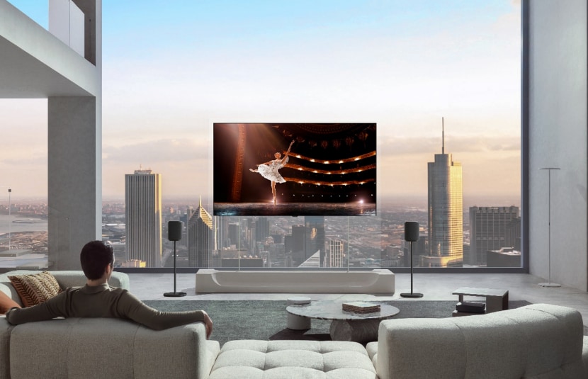A man is sitting on a sofa in his city apartment, watching a ballerina performance on an LG OLED evo AI W6 Wallpaper TV mounted on a transparent stand with two speakers.