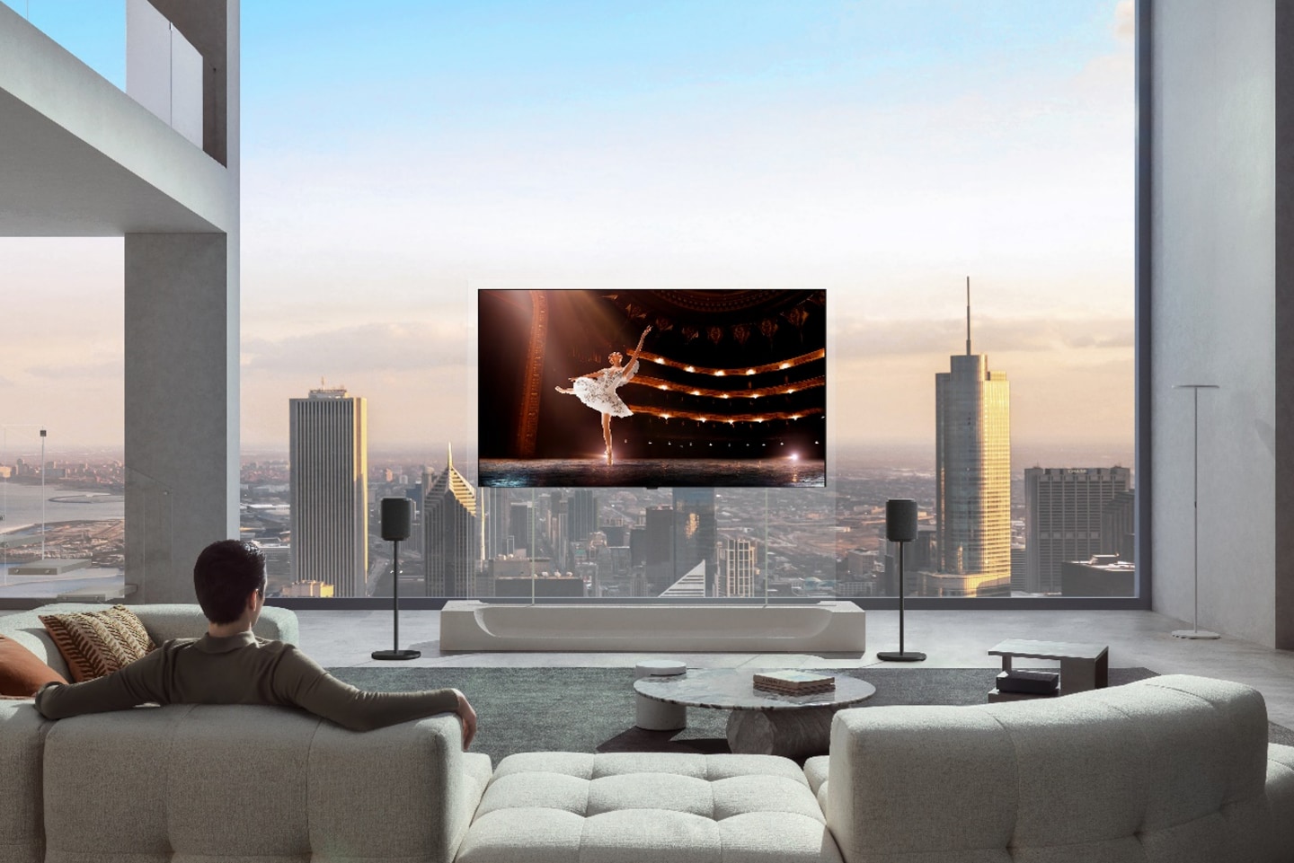 A man is sitting on a sofa in his city apartment, watching a ballerina performance on an LG OLED evo AI W6 Wallpaper TV mounted on a transparent stand with two speakers.