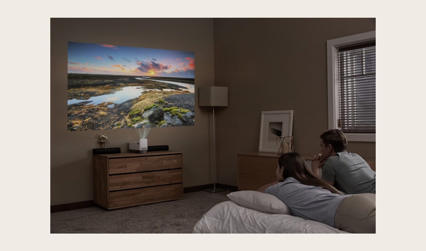 A couple lying in bed while watching a movie projected on the wall by the LG ProBeam Projector HF85J