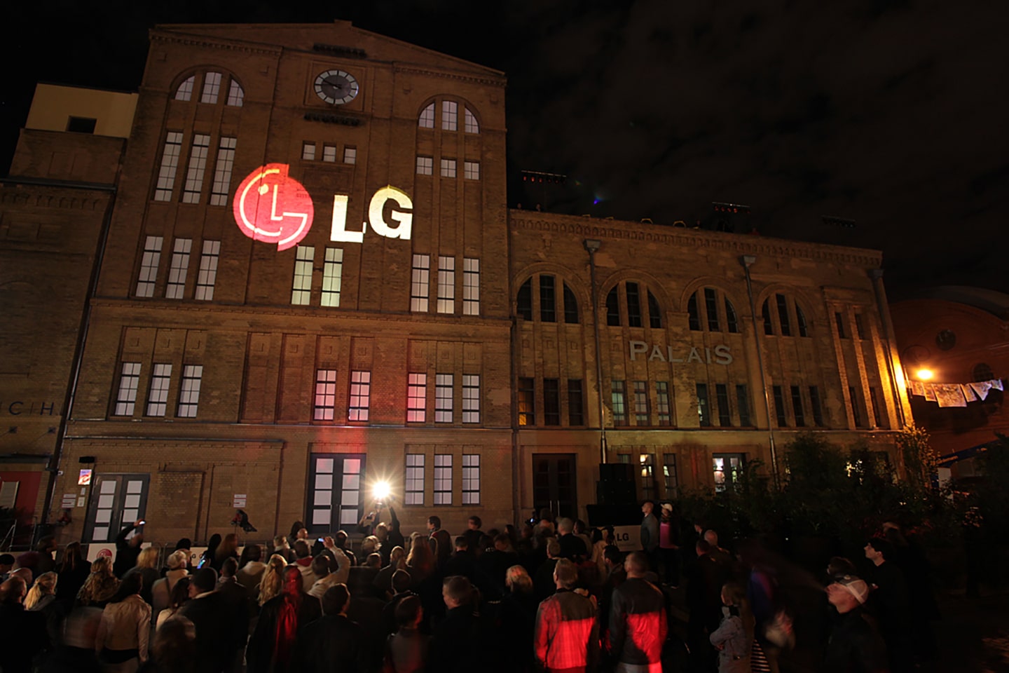 A large audience observes a gigantic 3D LG logo projected onto the front of a building in Kulturbrauerei, Berlin.