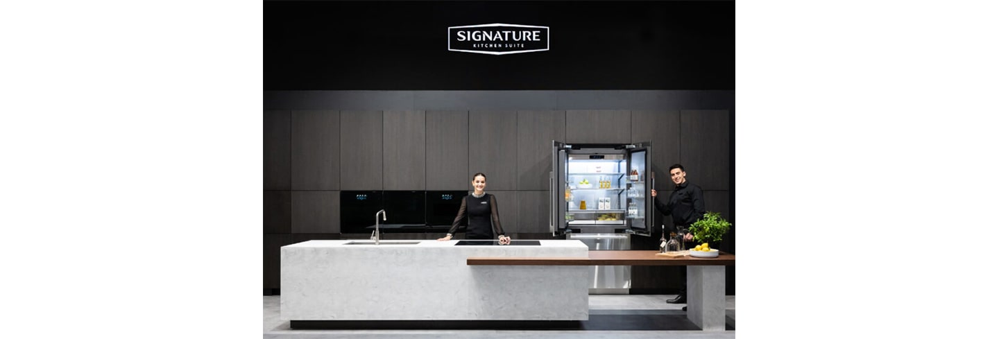 A woman is standing behind the kitchen island and a man is holding the Signature Kitchen Suite built-in 36-inch French Door refrigerator door at LG booth