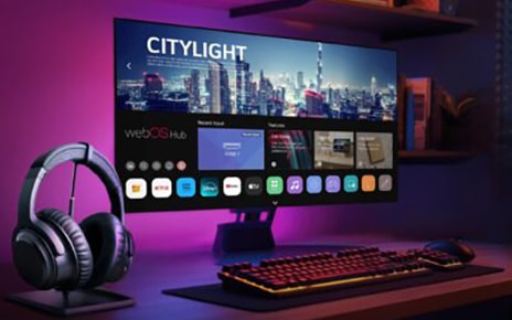 The monitor of a gaming setup displays the webOS Hub platform, with wireless headphones placed beside the keyboard