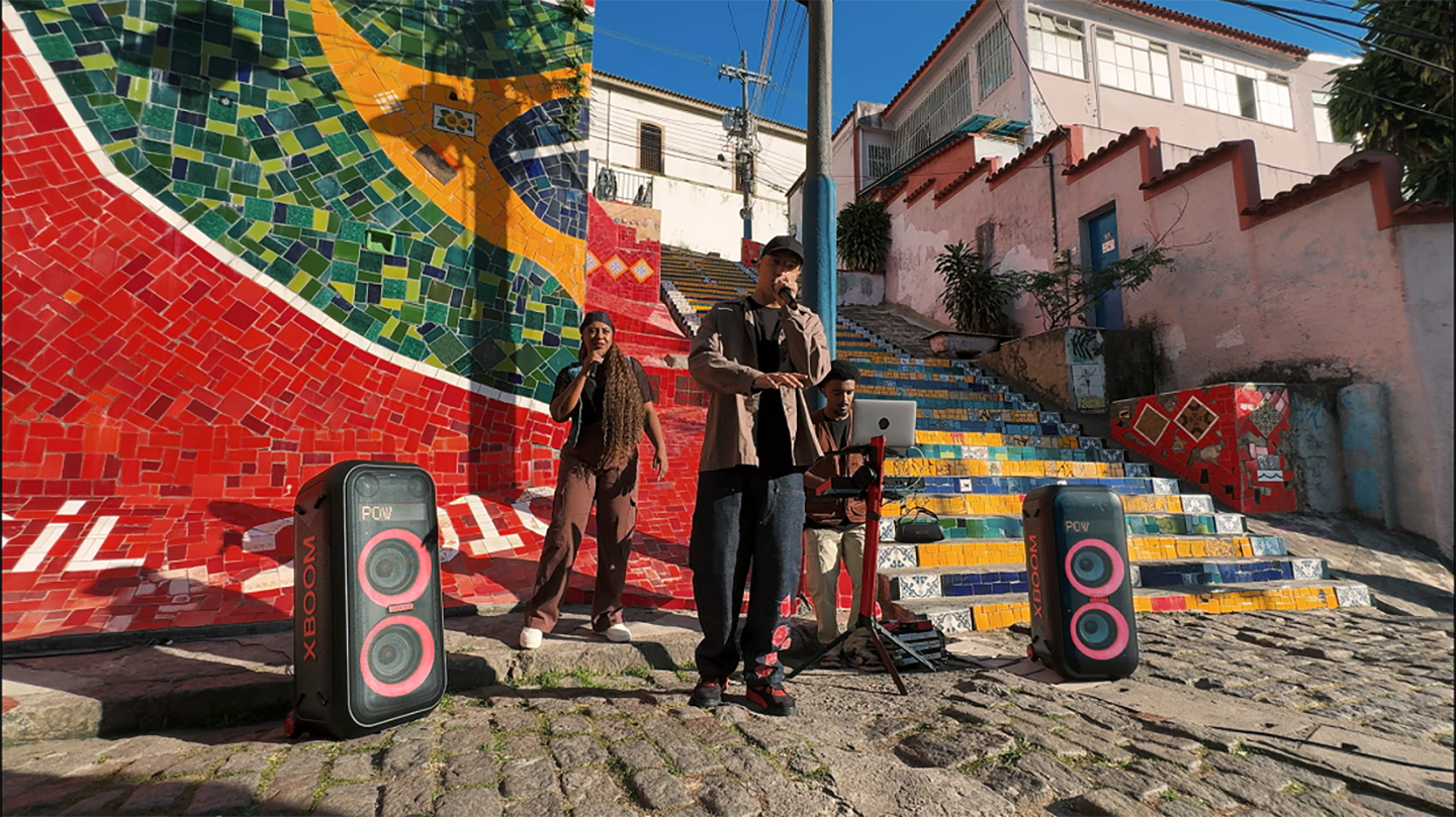 A group of three street performers in Brazil deliver a vibrant performance in front of a colorful mosaic mural and staircase, accompanied by two LG XBOOM speakers