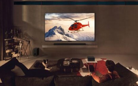 A man is watching a helicopter on the screen of a wall-mounted LG OLED M4, with a wireless Zero Connect Box sitting on a small table in the living space