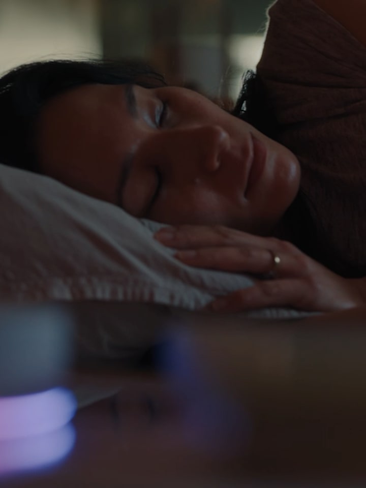 A woman lies on her side, sleeping in bed. In front of her, the LG ThinQ ON device is glowing. 