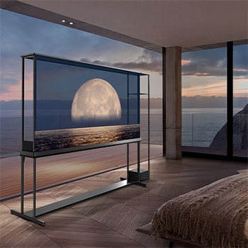 LG SIGNATURE OLED T 在透明螢幕上顯示描繪在海岸上方的月亮。LG SIGNATURE OLED T 置於房間中央，可看到懸崖邊的海景。右邊是一張床和一張躺椅。