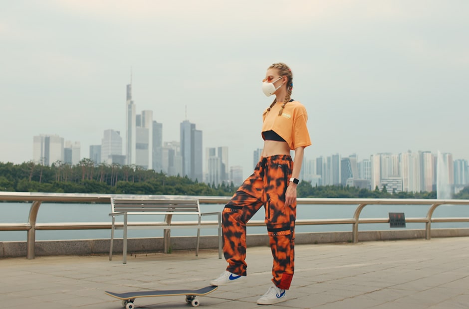 A young woman with braided hair puts her right foot on a skateboard, with a white LG PuriCare™ Wearable Air Purifier on.