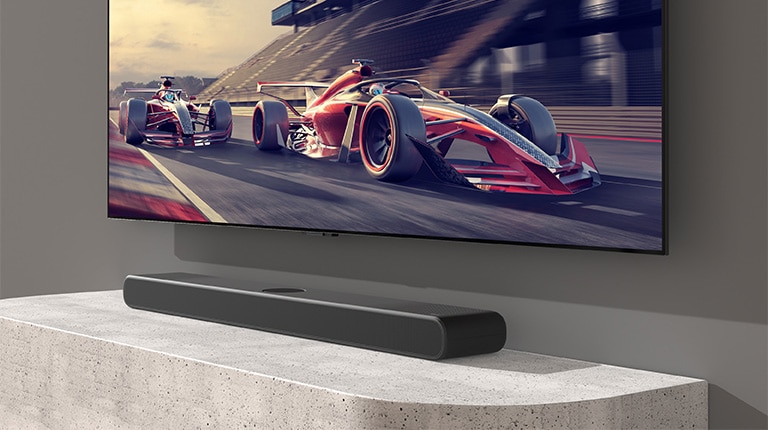 Sound bar and TV are placed on the white table and a racing scene is shown on the TV.