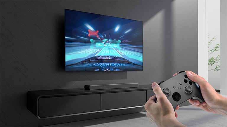 The sound bar is placed on the cabinet and racing game scene is shown on the TV coneected to the sound bar. A game console is on the bottom right side of the picture hold by two hands.
