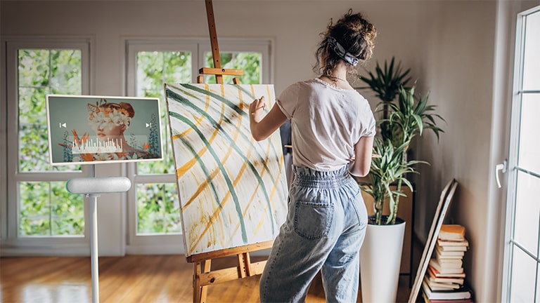 The LG StanbyME is placed in the livingroom with the LG StanbyME Speaker XT7S attached. A woman draws while listening to music through the screen and speaker combo.