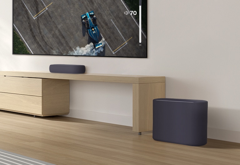 A soundbar is placed on a wooden table, a subwoofer is placed on a wooden floor. A mild sound wave effect comes out from the subwoofer on a floor. There is a TV placed above a soundbar. A racing car on a TV screen is driving very fast and violently and the GUI of TV volume is going up.
