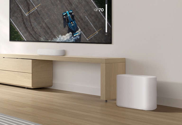 A soundbar is placed on a wooden table, a subwoofer is placed on a wooden floor. A mild sound wave effect comes out from the subwoofer on a floor. There is a TV placed above a soundbar. A racing car on a TV screen is driving very fast and violently and the GUI of TV volume is going up.