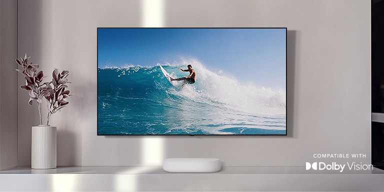 TV is on the wall. TV shows a man surfing on big wave. LG Soundbar is right below TV on a white shelf. There is a vase with a flower right next to the soundbar.Dolby Vision logo placed on right bottom corner.