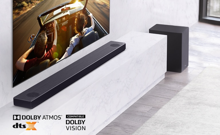 TV is on the wall, LG Soundbar is below on a white marble shelf with a sub-woofer to the right. TV shows a couple in a car.