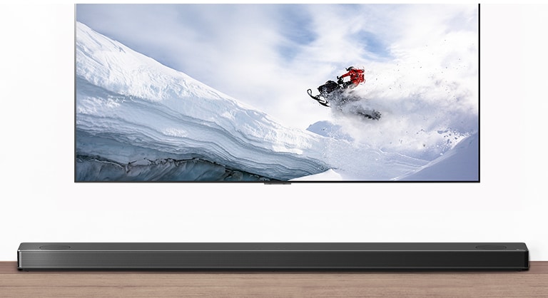 TV and Soundbar are seen from the front. TV shows man riding snowmobile in the snowy mountains. HDMI 2.1 logo is below TV.