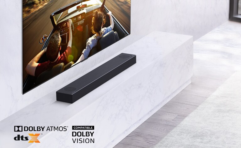 TV is on the wall, LG Soundbar is below on a white marble shelf with a sub-woofer to the right. TV shows a couple in a car.