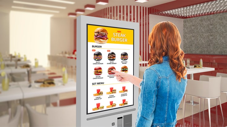 A woman is touching a kiosk to select a hamburger from the menu.