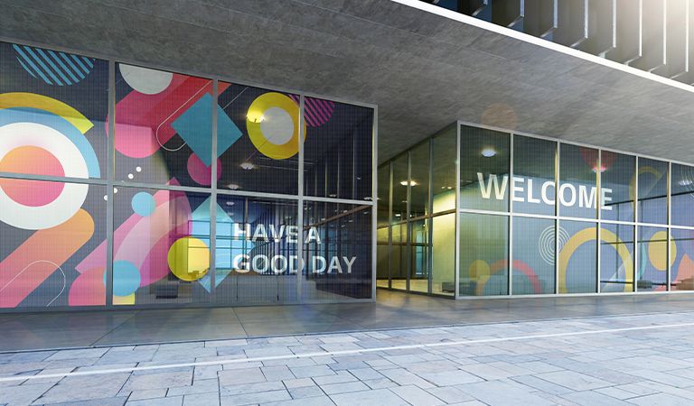 The transparent LED films installed on the glass wall at the building's entrance show content welcoming visitors. Some of the content shows the color well, and some of it looks transparent enough for the background behind the LED films to be seen.