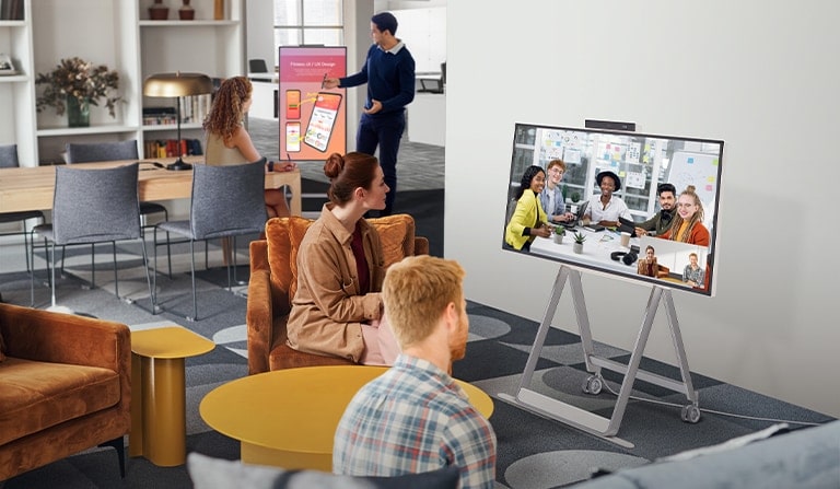 Two people are having a video conference using One:Quick Flex in a space with an informal atmosphere. The other two people at another table behind them are having a conference while flipping One:Quick Flex vertically and marking or writing on it with a dedicated touch pen on the screen.