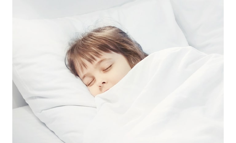 Person with light brown hair lying on a white pillow, covered by a white blanket, resting peacefully in bed.