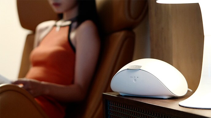 A woman in an off-the-shoulder dress is sitting on an armchair, and the PuriCare wearable case is placed on a small chest of drawers.