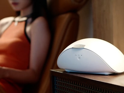 A woman in an off-the-shoulder dress is sitting on an armchair, and the PuriCare wearable case is placed on a small chest of drawers.