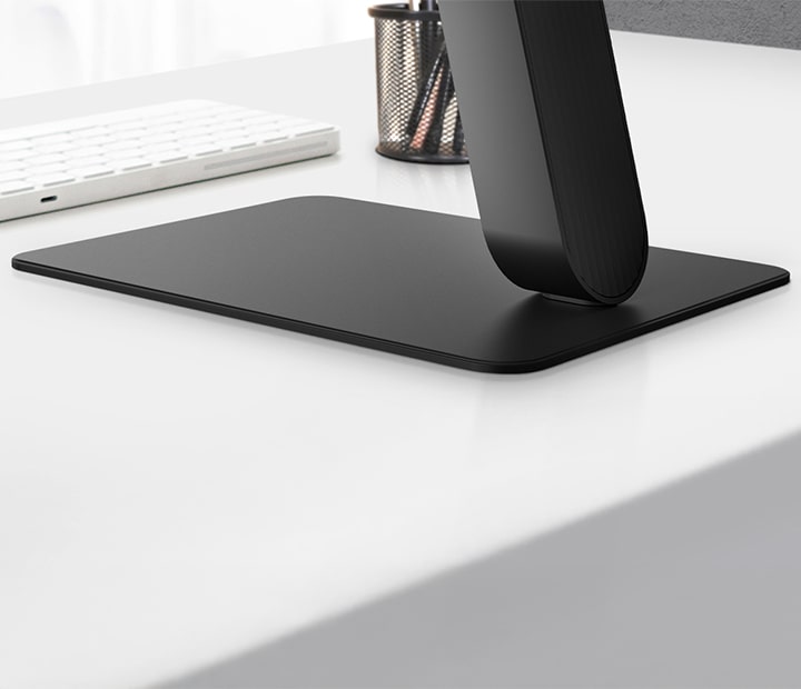 A close-up view of a sleek black monitor stand with a flat rectangular base, placed on a white desk beside a white keyboard and pen holder. The minimalist design emphasizes stability and elegance, blending seamlessly into a modern workspace.