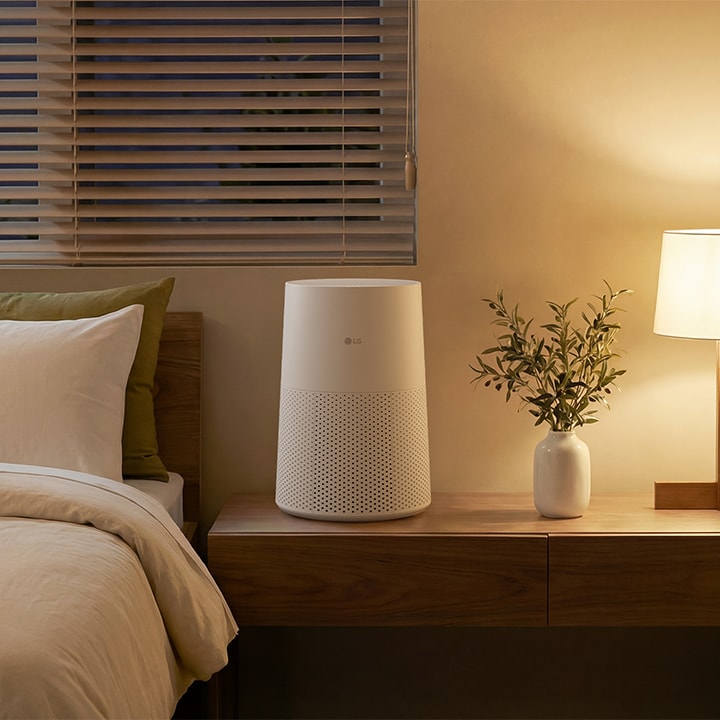 A white LG air purifier on a wooden bedside table next to a glowing warm lamp, a small plant, and a bed in a cozy, dimly lit bedroom	