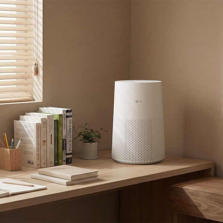 A white LG air purifier on a wooden desk in a bright room with sunlight filtering through blinds, next to books, a tablet, and a small plant	