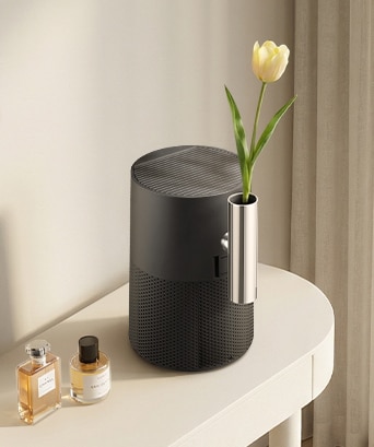 A matte black LG air purifier on a white vanity, featuring a sleek silver vase attachment with a single yellow tulip next to perfume bottles.	