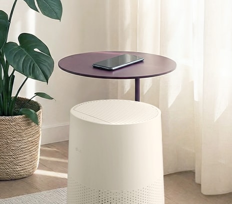 A cream-colored LG air purifier integrated with a round purple side table, holding a smartphone in a sunlit room near a large potted plant	